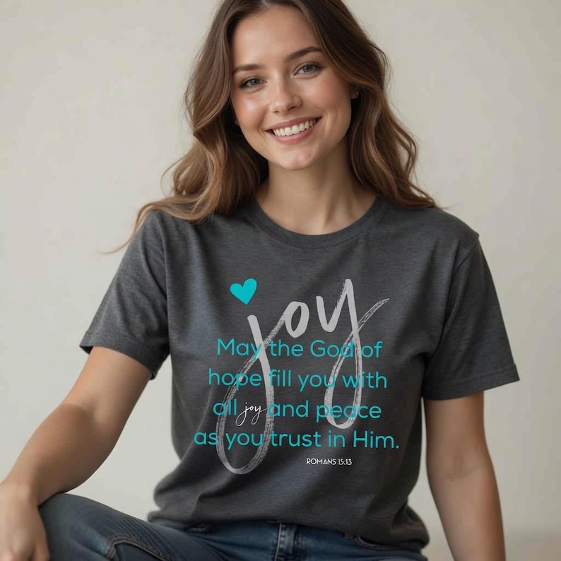 May include: Dark gray t-shirt with the word "joy" in white script and a teal heart. The shirt features the text "May the God of hope fill you with all joy and peace as you trust in Him." from Romans 15:13.