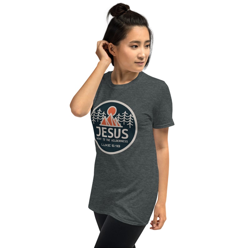 May include: A dark heather grey t-shirt with a circular graphic design featuring a mountain range, a sun, and pine trees. The graphic also includes the text "JESUS WENT TO THE WILDERNESS LUKE 5:16-18".