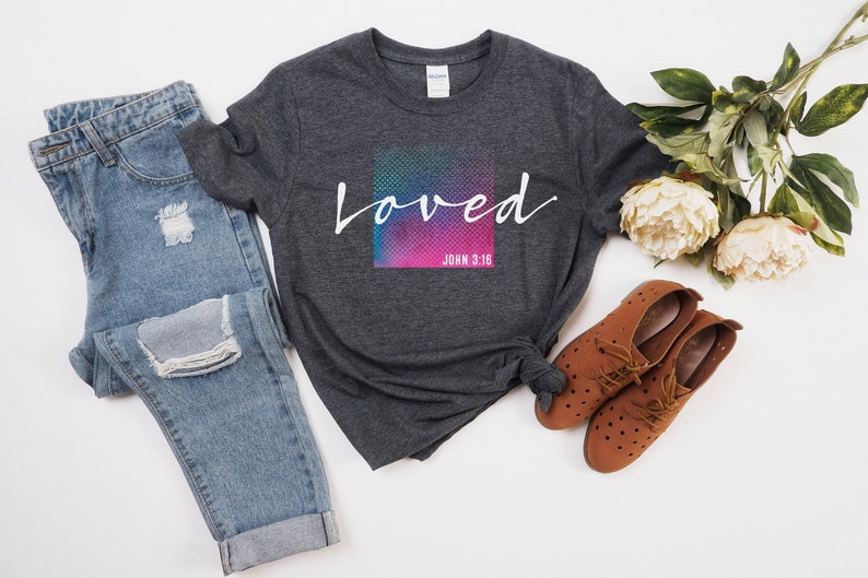 Pu&ograve; includere: Una maglietta grigia scura con una grafica bianca che dice "loved" con uno sfondo colorato e il testo "John 3:16" sotto. La maglietta &egrave; abbinata a jeans blu e scarpe marroni.