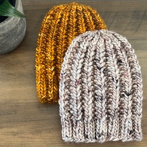 May include: Two knitted hats, one in a mustard yellow color and the other in a light brown with white flecks. Both hats have a ribbed texture.
