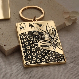 Può includere: Portachiavi in metallo dorato con un'illustrazione in bianco e nero di un giaguaro con un motivo floreale. Il portachiavi ha un anello per attaccare le chiavi.