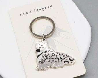 May include: A silver-colored snow leopard keychain with black spots and details, attached to a metal ring. The keychain is on a cream-colored card with the words "snow leopard" printed on it.