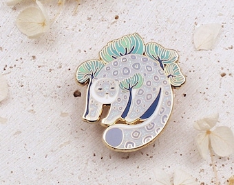 May include: A gold-tone enamel pin featuring a white snow leopard with black spots, surrounded by green and blue trees. The pin is on a white textured background.