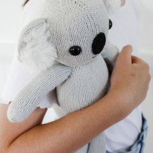 Könnte beinhalten: Ein gestricktes graues Koala-Plüschtier mit schwarzer Nase und Augen sowie weißen, flauschigen Ohrakzenten. Der Koala wird gehalten, was seine weiche Textur und detaillierte Verarbeitung zeigt. Ein bezauberndes und kuscheliges Stofftier.