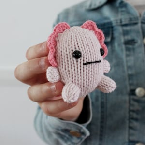 May include: A pink knitted axolotl plush toy with black eyes and a black mouth. The toy has pink frills around its head and is being held by a person.