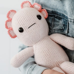 May include: A pink knitted axolotl plush toy with black eyes and pink frills around its head. The toy is being held by a person wearing a denim jacket.