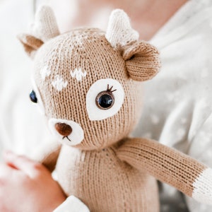 Peut inclure: Un jouet en peluche en forme de cerf en tricot marron avec des accents blancs et de grands yeux noirs. Le jouet est tenu par une personne.