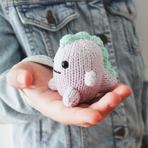 May include: A small, knitted purple dinosaur toy with a green spiky back and black eyes. The dinosaur is being held in the palm of a hand.