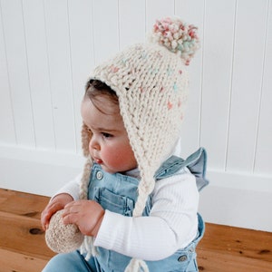 May include: A white knitted baby hat with a pom-pom on top and ear flaps. The hat has a colorful star pattern and is worn by a baby wearing a blue denim overall.