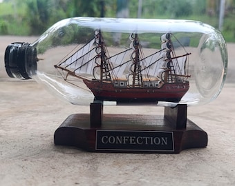 May include: A miniature wooden sailing ship inside a clear glass bottle. The ship has three masts with white sails and a red hull. The bottle sits on a dark wooden base with the word "CONFECTION" on a small plaque.