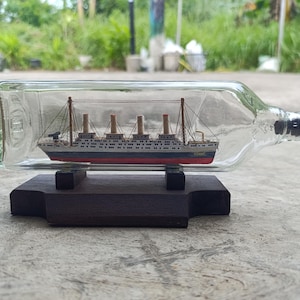 Könnte beinhalten: Eine klare Glasflasche mit einem Miniatur-Schiffsmodell. Das Schiff ist weiß, rot und schwarz, mit vier Schornsteinen und Takelage. Die Flasche steht auf einem dunkelbraunen Holzständer, der ein maritimes Thema zeigt.