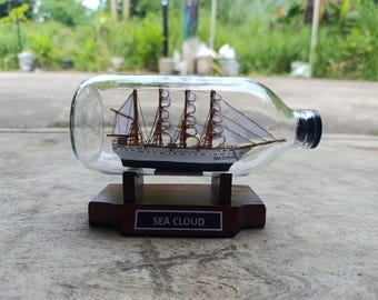 May include: A miniature ship model of the Sea Cloud, a white sailing ship with brown masts and sails, is displayed inside a clear glass bottle on a wooden base. The base has a plaque that reads "SEA CLOUD".