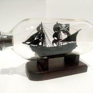 May include: A black pirate ship model in a clear glass bottle with a wooden base. The ship has a skull and crossbones flag.