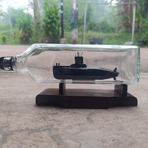 May include: A clear glass bottle containing a small black submarine model, resting horizontally on a dark brown wooden stand. The submarine features a conning tower with visible periscopes and a propeller at the rear, all enclosed within the bottle.