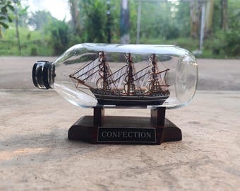 May include: A miniature wooden ship with white sails inside a clear glass bottle. The bottle is mounted on a dark wooden base with the word "CONFECTION" on a small plaque. The ship has three masts and is painted black and brown.