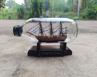 May include: A miniature wooden sailing ship inside a clear glass bottle. The ship has three masts with white sails and is mounted on a wooden base. The base has a small plaque that reads "CONFECTION".