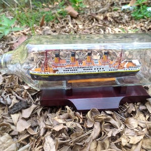 Könnte beinhalten: Ein Miniaturmodell der Titanic wird in einer durchsichtigen Glasflasche auf einem Holzsockel ausgestellt. Das Schiff ist mit orangefarbenen und braunen Details bemalt und der Schriftzug "TITANIC" ist an der Seite sichtbar.