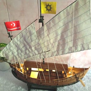 Peut inclure: Un modèle réduit de bateau en bois avec deux grandes voiles en tissu blanc cassé, un drapeau rouge avec un croissant et un sabre blancs, et un drapeau jaune avec un emblème de soleil noir. La coque est en bois brun avec éclairage intérieur.