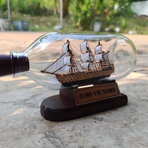 Puede incluir: Un detallado modelo de barco de madera dentro de una botella de vidrio transparente. El barco tiene velas blancas y está montado sobre una base de madera con el texto "HMS Victory". La botella tiene un corcho marrón oscuro.