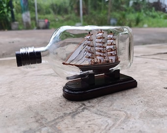 May include: A miniature wooden sailboat inside a clear glass bottle. The ship has white sails and brown masts, set on a dark wooden base. The bottle has a black cap and is displayed on a concrete surface.