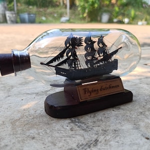 May include: A clear glass bottle with a dark brown cork and a miniature black sailing ship inside. The ship is mounted on a wooden base with a plaque reading "Flying Dutchman". The bottle is displayed on a concrete surface.