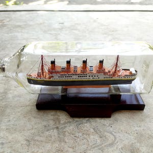 May include: A miniature model of the Titanic ship inside a clear glass bottle. The ship is made of paper and has a brown hull with white accents. The bottle is sitting on a wooden base.