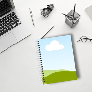 May include: A spiral-bound notebook with a landscape design featuring a blue sky, white cloud, and green hills. The notebook is open on a white desk, surrounded by a laptop, pens, and other office supplies.