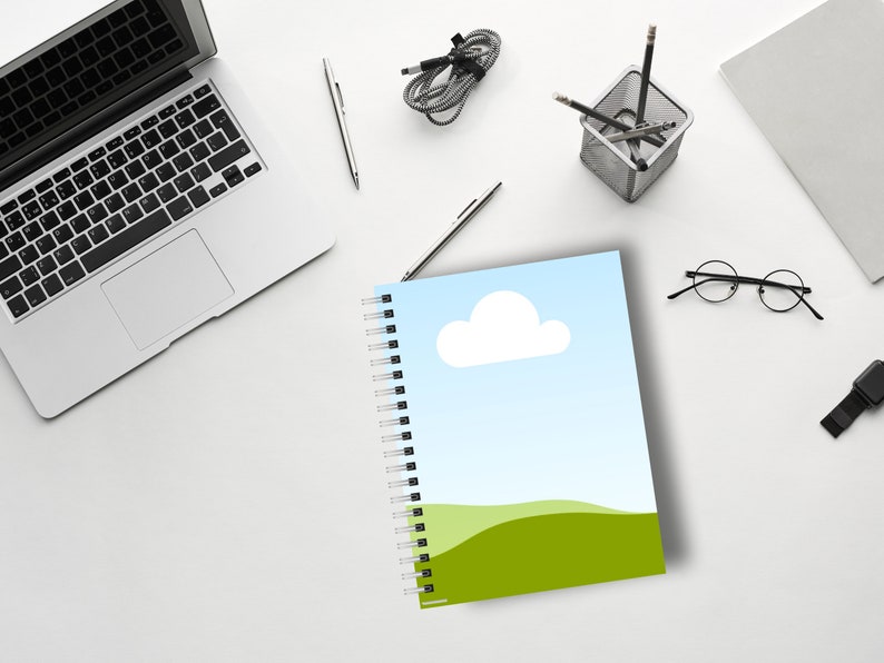 May include: A spiral-bound notebook with a landscape design featuring a blue sky, white cloud, and green hills. The notebook is open on a white desk, surrounded by a laptop, pens, and other office supplies.