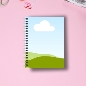 May include: A spiral-bound notebook with a cover featuring a blue sky, white cloud, and green hills design. The notebook is open, revealing the spiral binding and blank pages. The background is a pink surface with office supplies.