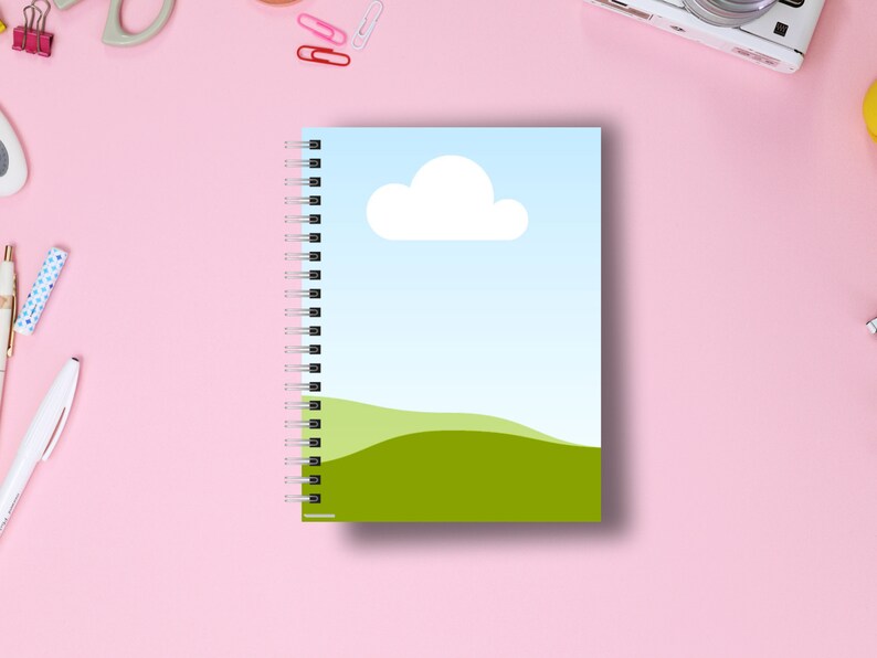 May include: A spiral-bound notebook with a cover featuring a blue sky, white cloud, and green hills design. The notebook is open, revealing the spiral binding and blank pages. The background is a pink surface with office supplies.