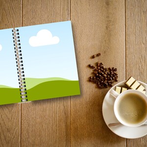 May include: An open spiral-bound notebook with a landscape illustration of green hills, blue sky, and white clouds. A cup of coffee, a saucer, a spoon, and pieces of chocolate are on a wooden surface. Coffee beans are scattered nearby.