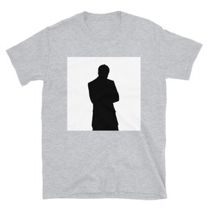 Puede incluir: Una camiseta gris con un cuadrado blanco que presenta una silueta negra de una persona de pie con los brazos cruzados.