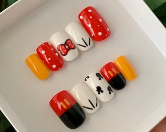 Könnte beinhalten: Satz mit sechs falschen Fingernägeln mit Disney-Motiv. Die Nägel sind weiß mit roten, gelben und schwarzen Akzenten. Die Designs beinhalten Minnies charakteristisches rotes Schleifenband, Punkte und Mickeys Kopf.
