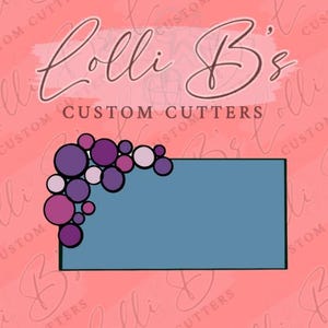 May include: A blue rectangle with a white border, surrounded by a cluster of purple, pink, and white circles with black outlines. The text "Lolli B's CUSTOM CUTTERS" is above the rectangle.