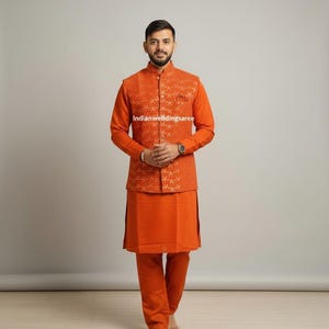 May include: An orange three-piece suit. The outfit includes a long-sleeved shirt, a vest with a geometric pattern, and matching pants. The suit is a solid orange color. The text "Indianweddingsaree" is visible.
