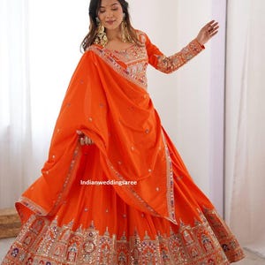 Peut inclure: Un ensemble lehenga orange vif composé d'un chemisier à manches longues et d'une jupe longue, tous deux ornés de broderies multicolores complexes, de miroirs et de détails métalliques. Un dupatta orange assorti est drapé sur l'ensemble.