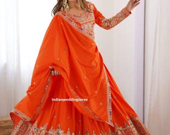 May include: An orange traditional Indian dress with intricate embroidery and embellishments. The dress features a long skirt, a fitted bodice, and a matching scarf. The sleeves and skirt hem are adorned with detailed patterns. The text "indianweddingsaree" is visible on the dress.
