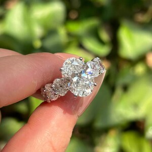 May include: A silver-toned ring featuring a large, central oval-cut gemstone flanked by smaller, round-cut stones along the band. The setting is metallic, and the stones are clear and faceted, reflecting light with a brilliant, sparkling appearance.