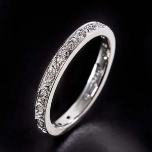 Puede incluir: Un anillo de plata con intrincados patrones florales y de hojas, engastado con pequeñas y brillantes gemas. La banda del anillo presenta grabados detallados, creando un diseño delicado y elegante. El anillo se muestra sobre un fondo oscuro.