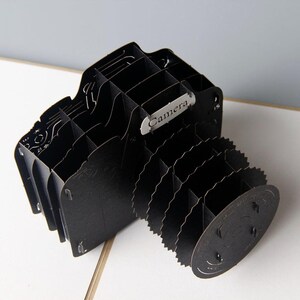May include: Black cardboard camera shaped desk organizer with multiple compartments. The camera has a silver label that reads "Camera".