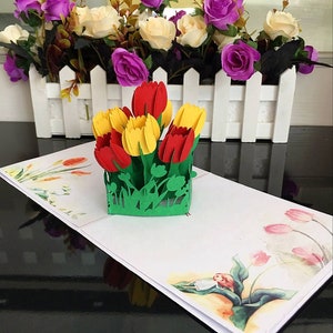 May include: A 3D pop-up card featuring a bouquet of red and yellow tulips. The card is open and the tulips are standing up in a green base. The card is on a white surface with a watercolor design of flowers and a fairy.