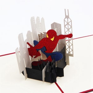 May include: A 3D pop-up card featuring Spiderman in a red and blue suit, swinging through a cityscape. The card is made of paper and has a black base.