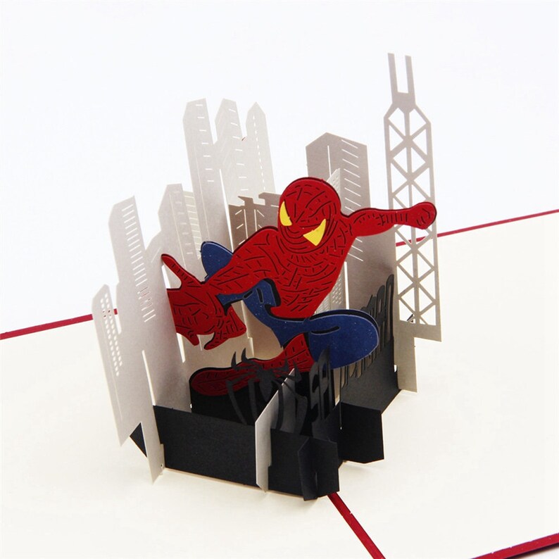 May include: A 3D pop-up card featuring Spiderman in a red and blue suit, swinging through a cityscape. The card is made of paper and has a black base.