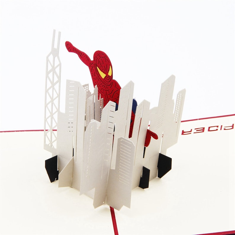 May include: A red and blue 3D pop-up card featuring Spiderman swinging through a cityscape. The card is open and the pop-up is fully extended. The card has the word "Spider" printed on the front.