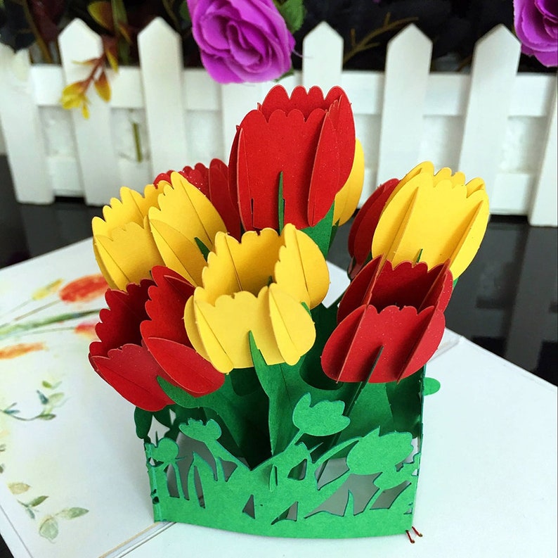 May include: A 3D pop-up card featuring a bouquet of red and yellow tulips with green leaves and stems. The tulips are arranged in a vase-like shape with a cut-out design.