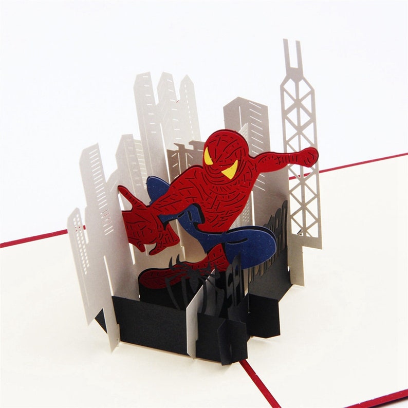 May include: A 3D pop-up card featuring Spiderman in a red and blue suit, swinging through a cityscape. The card is made of paper and has a black base.