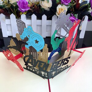 May include: A 3D pop-up card featuring the Avengers. The card is open and shows the characters Captain America, Hulk, Iron Man, and Thor. The card is made of paper and has a gold and black color scheme. The card says "Avengers" in black letters.