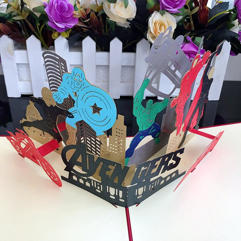 May include: A 3D pop-up card featuring the Avengers. The card is open and shows the characters Captain America, Hulk, Iron Man, and Thor. The card is made of paper and has a gold and black color scheme. The card says "Avengers" in black letters.