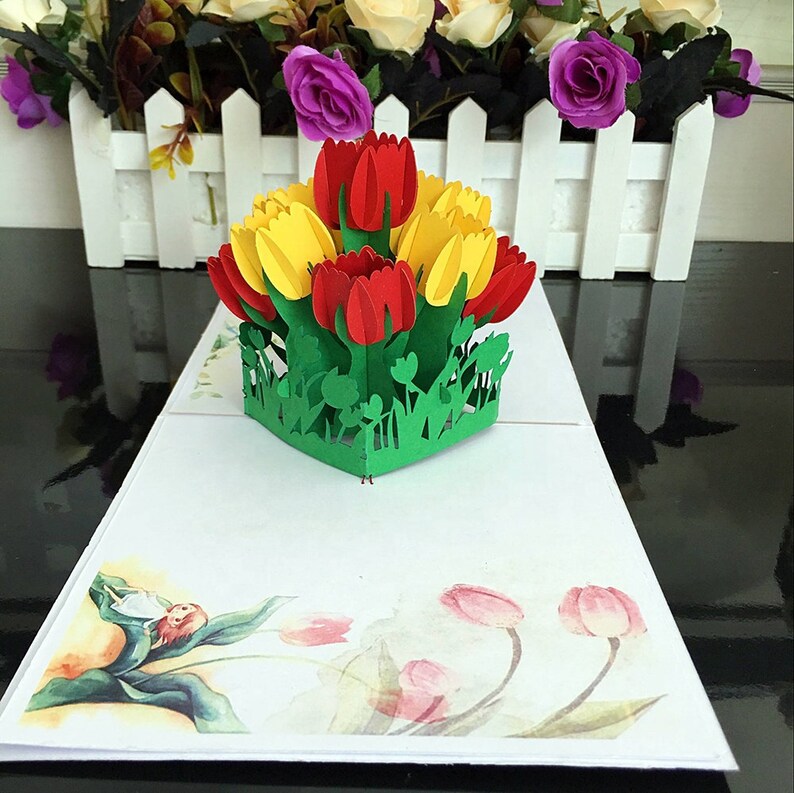 May include: A 3D pop-up card featuring a bouquet of red and yellow tulips with green leaves. The card is open and the tulips are standing up. The background of the card is white with a watercolor design of pink tulips.