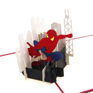 May include: A red and blue 3D pop-up card featuring Spiderman swinging through a cityscape. The card is made of paper and has a black base.